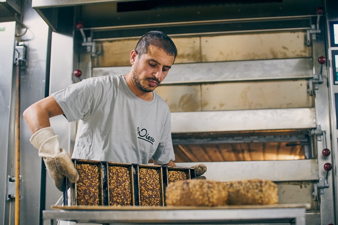 Ein Mann, der Brotlaibe aus einem Backofen holt. Klick führt zu Großansicht.