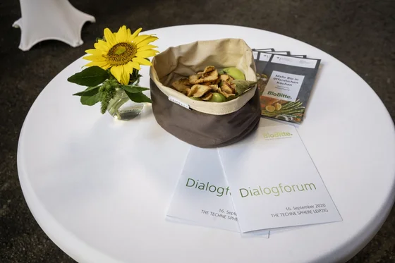 Stehtisch mit Sonnenblume in Vase, Korb mit Äpfeln und Chips sowie Infomaterialien. Klick führt zu Großansicht in einer Lightbox. Zum Schließen der Lightbox ESC drücken.