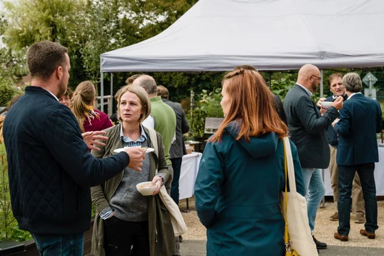 Menschen unterhalten sich in kleiner Gruppe bei einem Outdoor-Event mit Zelt, einige essen oder trinken, entspannte Atmosphäre. Klick führt zu Großansicht in einer Lightbox. Zum Schließen der Lightbox ESC drücken.