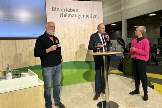 eine Frau unterhält sich auf der Bühne mit zwei Männern die Mikrofone in der Hand halten.