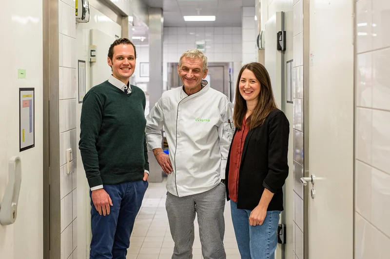 Tim Marx, Tassilo Springer und Alina Koch stehen lächelnd in einem Flur der DI-VERSA-Großküche. Springer trägt eine weiße Kochjacke.