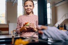 Mädchen mit Essen in der Hand. Sie lacht freundlich in die Kamera. Klick führt zu Großansicht.