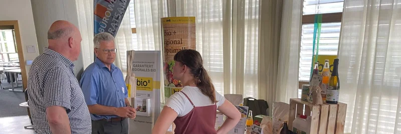 Drei Personen stehen in einem Veranstaltungsraum an einem BioBitte-Infostand mit regiona-len Bio-Produkten und Informationsmaterialien und sprechen miteinander.