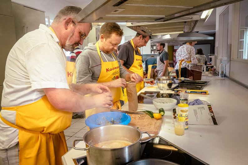 Drei Männer in gelben Kochschürzen stehen in der Küche und arbeiten auf der Arbeitsfläche