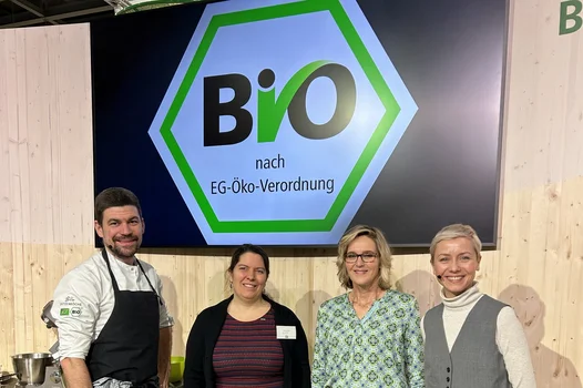 Ein Mann und drei Frauen stehen vor einem großen Bild des Bio-Siegels und lachen in die Kamera.
