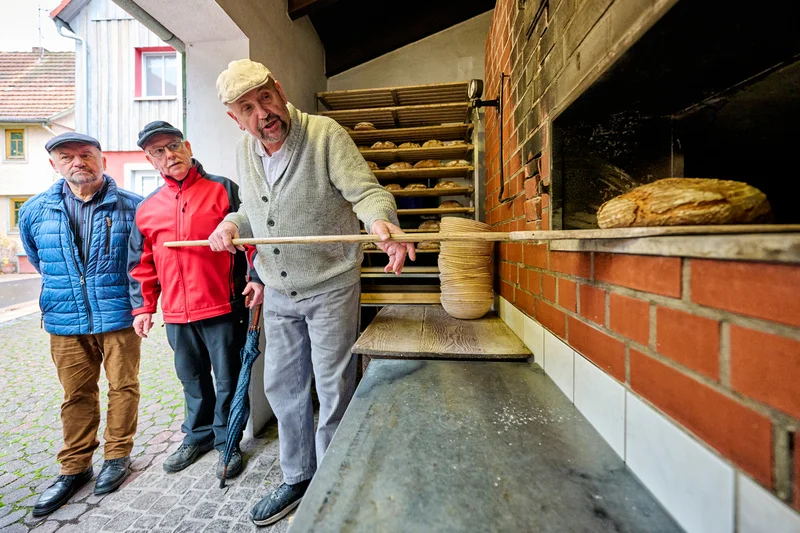 Drei Männer stehen vor einem traditionellen Backofen, während ein Mann ein Brot aus dem Ofen holt. Klick führt zu Großansicht in neuem Fenster.