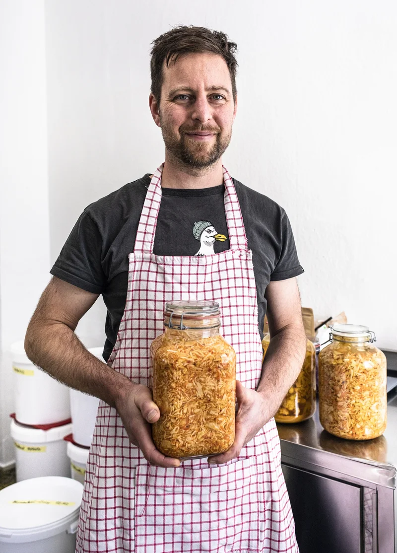 Ein Mann mit Schürze und einem Glas mit fermentiertem Gemüse in der Hand. Klick führt zu Großansicht.