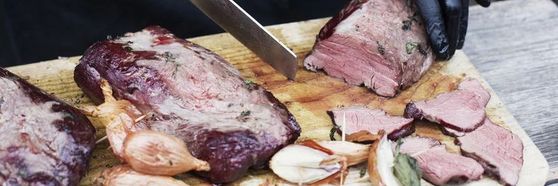 Zu sehen ist ein Stück Bratenfleisch auf einem Holzbrett. Ein Mensch schneidet das Stück in Scheiben.