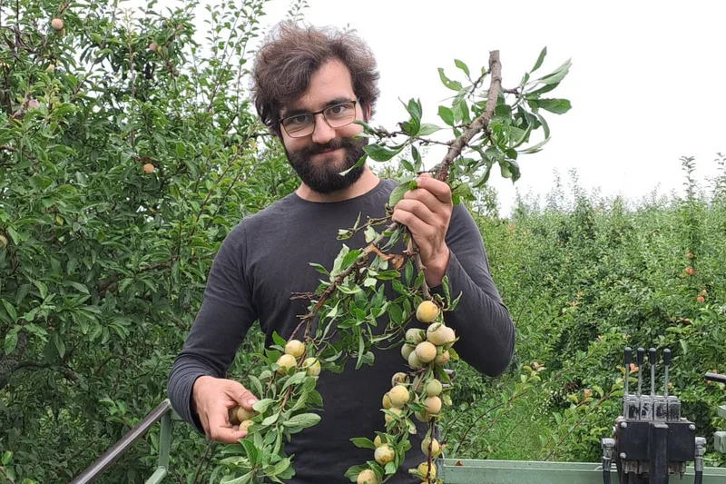 Junger Mann mit Obstbaumzweigen in der Hand blickt in die Kamera. Klick führt zu Großansicht.