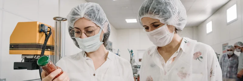 Zwei Frauen mit Laborkittel, Haarnetz und Maske bei einer Lebensmittelkontrolle.