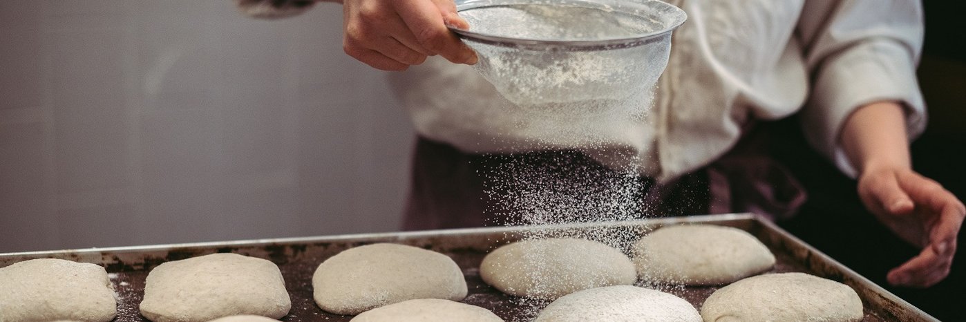 Eine Hand, wie sie Mehl über Teiglinge auf einem Backblech siebt.