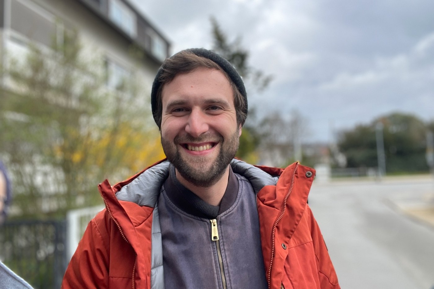 Ein Mann mit Mütze und roter Jacke lacht freundlich in die Kamera. Klick führt zu Großansicht.