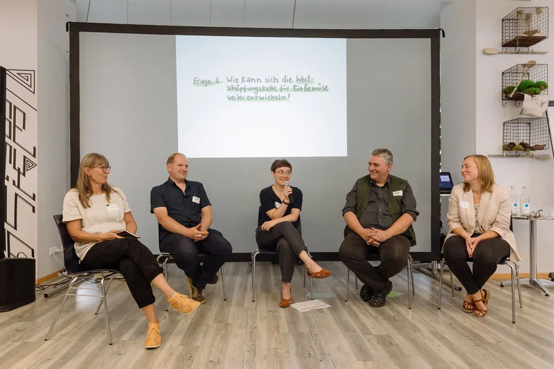 Fünf Menschen sitzen auf Stühlen nebeneinander. Klick führt zu Großansicht in einer Lightbox, Schließen per ESC.