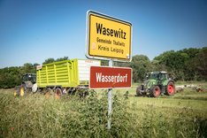 Im Vordergrund ist das Ortsschild von Wasewitz zu sehen sowie ein Schild mit der Aufschrift "Wasserdorf". Im Hintergrund Traktoren, die auf einem Acker Feldarbeiten verrichten. Klick führt zu Großansicht.