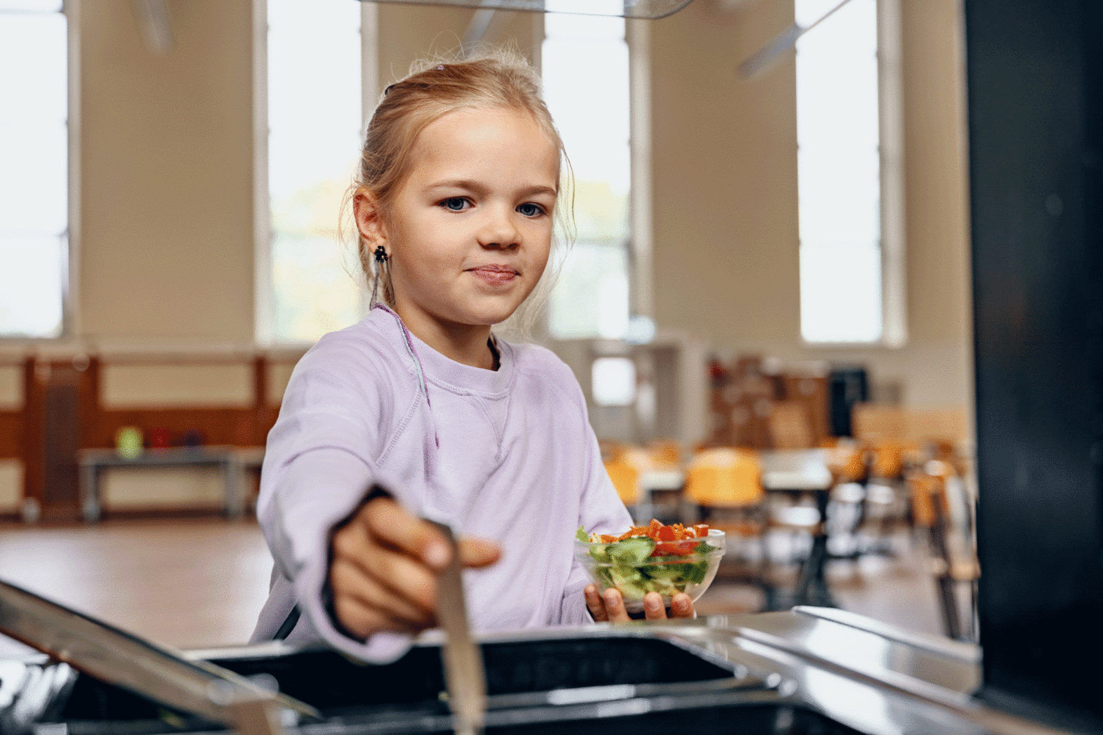 Mädchen an einer Essensausgabe mit Tablett und kleiner Salatschale in der Hand.