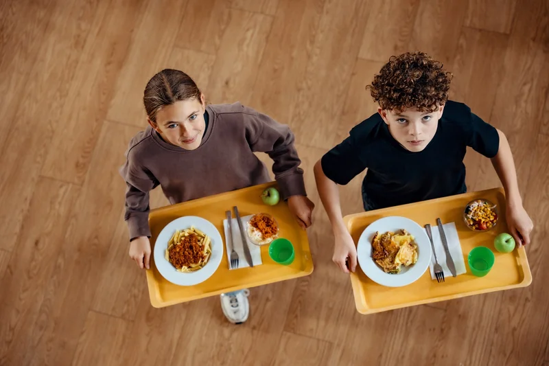 Zwei Kinder tragen Tabletts mit Tellergericht und grünem Becher und schauen nach oben; Aufnahme von oben auf einem Holzfußboden. Klick führt zu Großansicht in einer Lightbox. Zum Schließen der Lightbox ESC drücken.