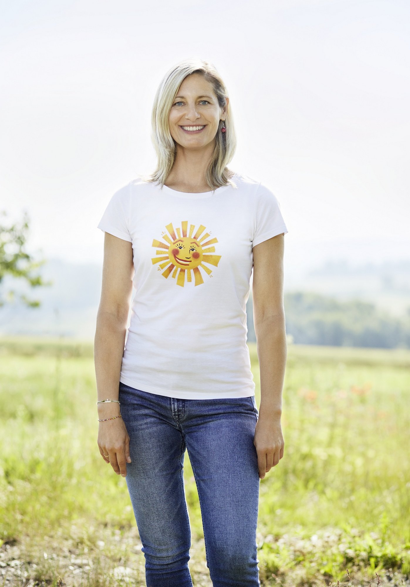 Frau mit weißem T-Shirt und einer Sonne darau steht auf einer Wiese und lacht in die Kamera. Klick führt zu Großansicht.