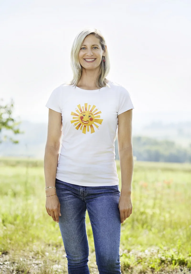 Frau mit weißem T-Shirt und einer Sonne darau steht auf einer Wiese und lacht in die Kamera. Klick führt zu Großansicht.
