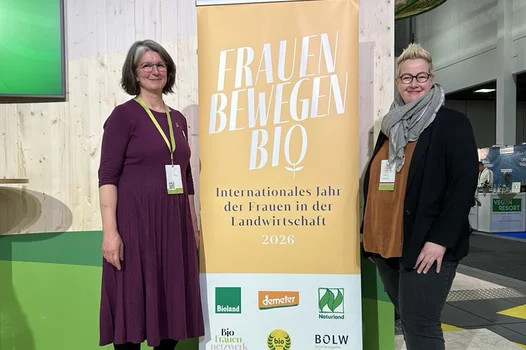 Zwei Frauen stehen neben einem Aufsteller auf dem "Frauen bewegen Bio" steht. Beide schauen freundlich in die Kamera.