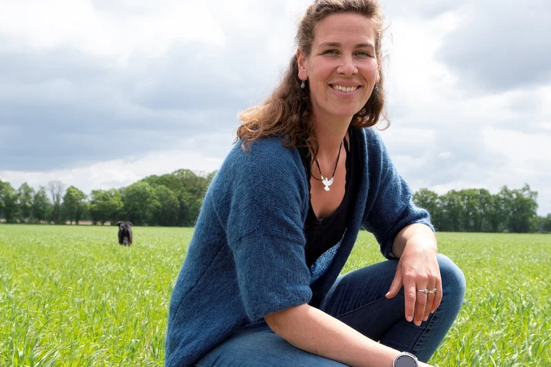 Frau hockt in einem Feld und lächelt in die Kamera.  (Klick führt zu Großansicht in einer Lightbox. Zum Schließen der Lightbox ESC drücken.) 