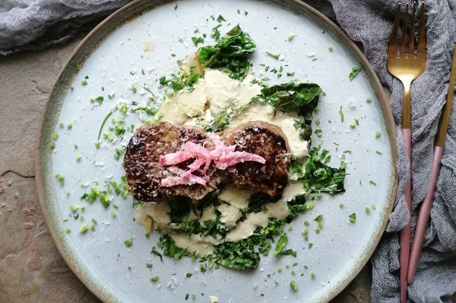 Trockengereiftes Rückensteak von der Backensholzer Färse mit lauwarmen Römersalat, Knoblauchemulsion und Deichkäse Gold auf einem Teller angerichtet.