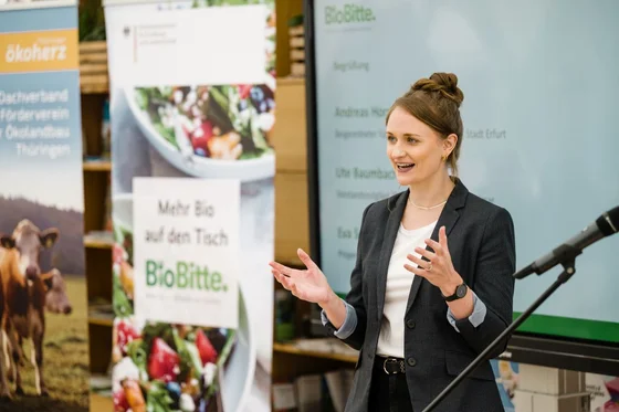 Frau spricht bei einer BioBitte-Veranstaltung vor Publikum, im Hintergrund Banner mit Bio-Motiven und Präsentationsfolie. Klick führt zu Großansicht in einer Lightbox. Zum Schließen der Lightbox ESC drücken.