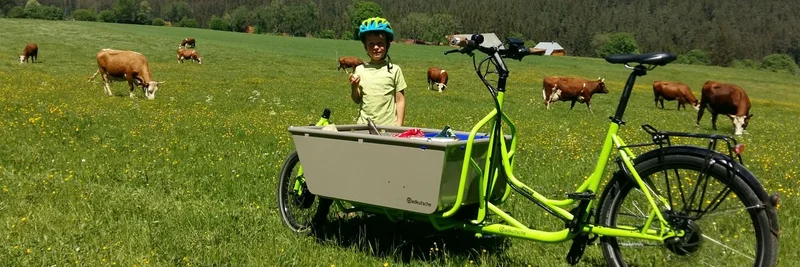 Kind steht hinter einem Lastenfahrrad auf einer Weide, im Hintergrund Kühe.