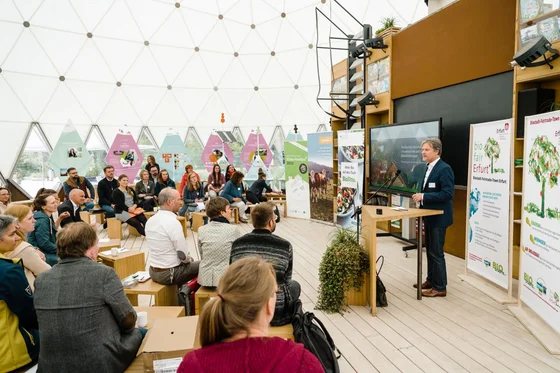 Mann spricht bei einer BioBitte-Veranstaltung vor aufmerksamem Publikum in einem hellen Raum mit Informationspostern und Holzmöbeln. Klick führt zu Großansicht in einer Lightbox. Zum Schließen der Lightbox ESC drücken.
