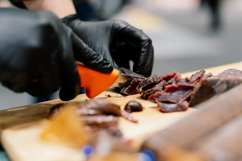 Hände wie sie Wurst auf einem Holzbrett aufschneiden. Klick führt zu Großansicht.