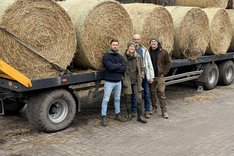 Vier Menschen, davon drei Männer und eine Frau, stehen vor einem Schlepper, der Strohballen geladen hat. Klick führt zu Großansicht.