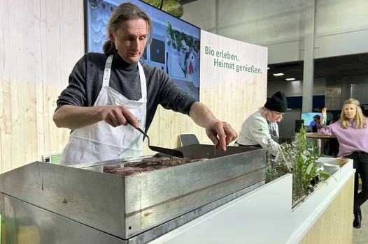 Ein Mann mit Kochschürze wendet Fleisch auf einem Kochfeld. Im Hintergrund sitzen zwei Frauen um einen Tisch und unterhalten sich.