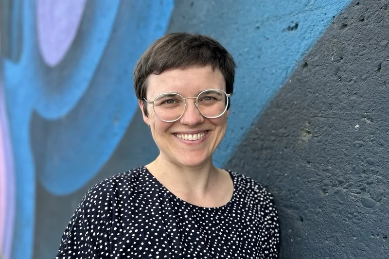 Eine Frau mit Brille steht vor einer blauen Wand und lächelt in die Kamera. Klick führt zu Großansicht.