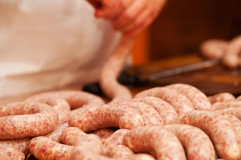 Der Prozess der Wurstherstellung ist dargestellt. Im Vordergrund liegen bereits fertig gefüllte Würste. Im Hintergrund sind Hände zu sehen, wie sie gerade Brät in eine Haut füllen. Klick führt zu Großansicht.