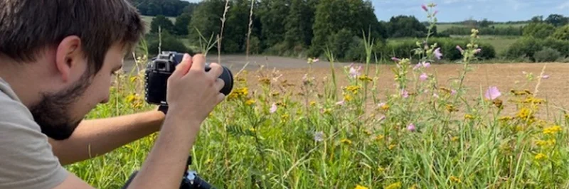 Mann hockt mit einer Kamera in einer Wiese.