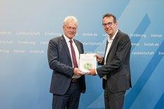 Zwei Männer halten eine Broschüre in der Hand und schauen in die Kamera. Klick führt zu Großansicht.