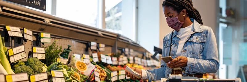 Frau geht Gemüse im Supermarkt einkaufen. In Anlehnung an die Corona-Krise trägt sie eine Maske.