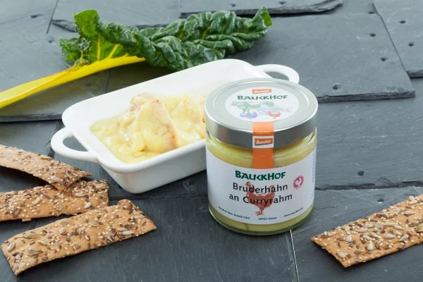 Ein Glas mit Hühnercurry, dahinter das Gericht angerichtet in einer Schale. Um die Auflaufform liegen Cracker und eine Stange Mangold zur Deko. Klick führt zu Großansicht.