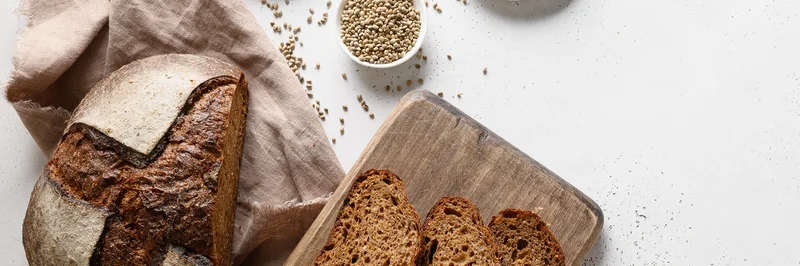Ein Brot, frisch angeschnitten. Auf einem Holzbrett liegen drei Scheiben Brot. Im Hintergrund sind Schälchen mit Mehl und Samen zu sehen.