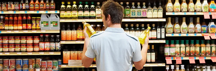 Junger Mann vergleicht zwei Flaschen Speiseöl im Supermarkt miteinander.