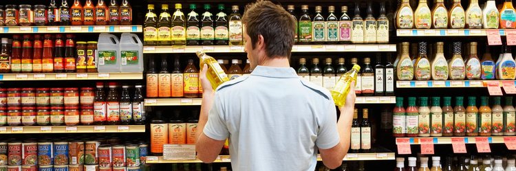 Junger Mann vergleicht zwei Flaschen Speiseöl im Supermarkt miteinander.