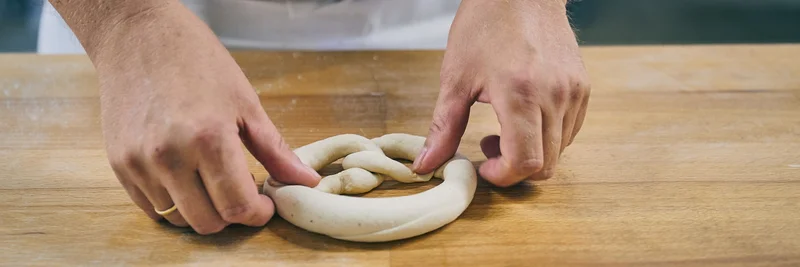 Hände, die einen Brezel-Teigling kneten.