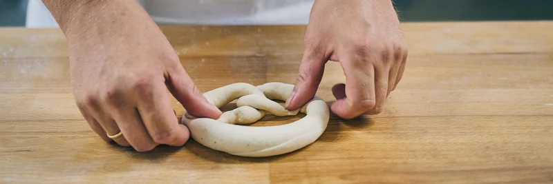 Hände, die einen Brezel-Teigling kneten.