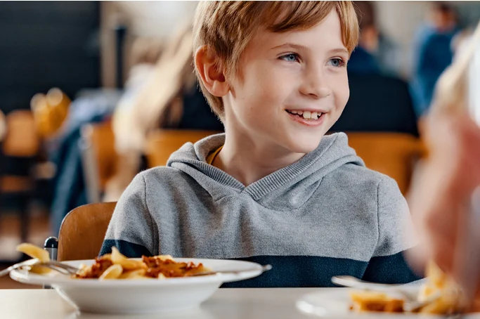 Ein Kind sitzt in einer Schulmensa vor einem Teller mit Essen und schaut zur Seite. Foto: BLE / BioBitte / Ilja Hendel
