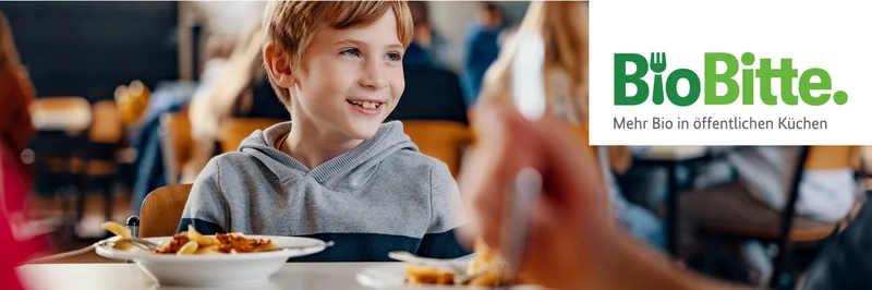 Ein Kind sitzt in einer Schulmensa vor einem Teller mit Essen und schaut zur Seite.