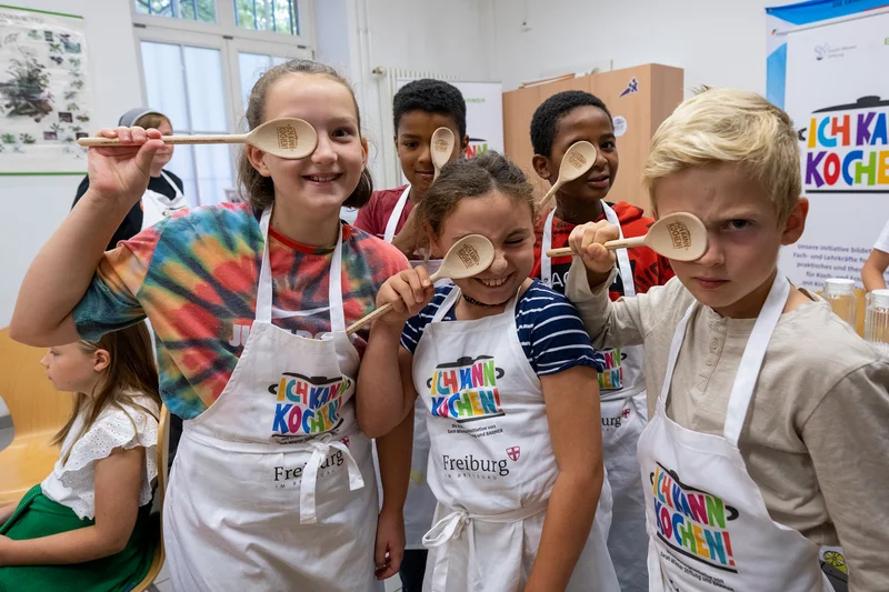 Schulkinder mit Schürze und Kochlöffel. Klick führt zu Großansicht in neuem Fenster.