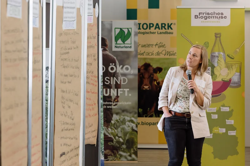 Frau mit Mikro in der Hand schaut auf Plakatwände. Klick führt zu Großansicht in Lighbtox, Schließen mit ESC.