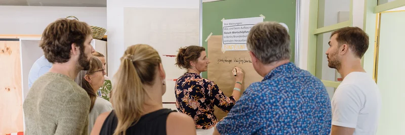 Eine Gruppe steht um eine Plakatwand, auf der eine Frau Notizen macht.