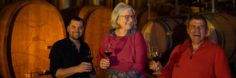 Zwei Männer und eine Frau in einem Weinkeller vor Weinfässern. Alle haben ein Glas Wein in der Hand.