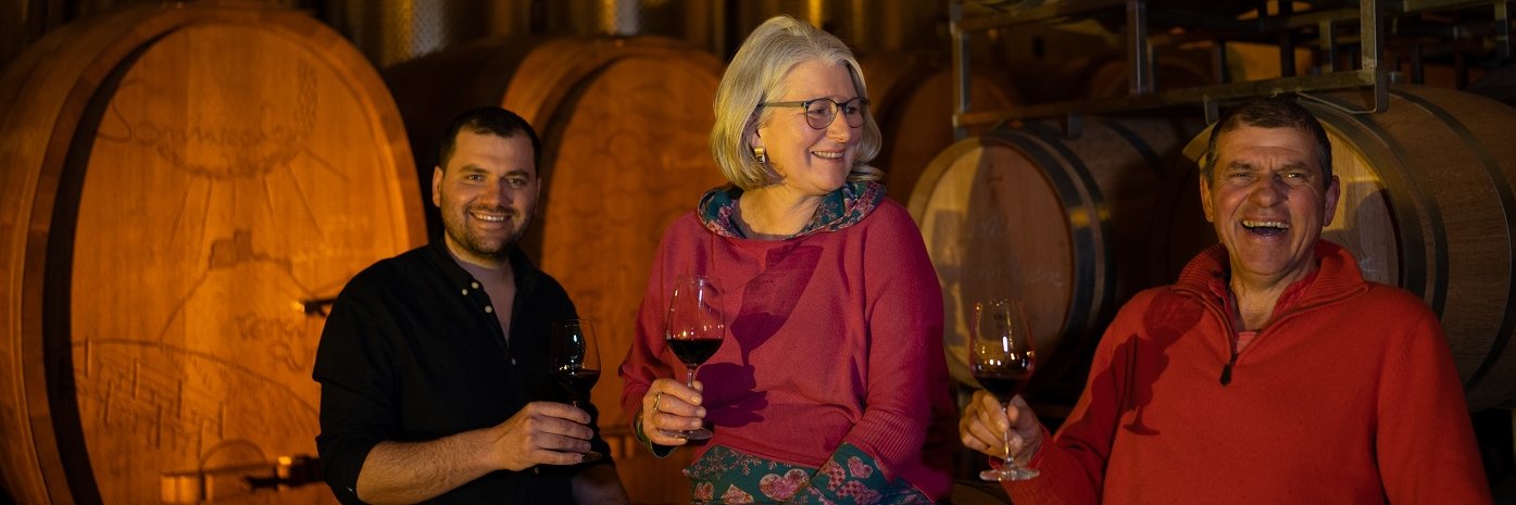 Zwei Männer und eine Frau in einem Weinkeller vor Weinfässern. Alle haben ein Glas Wein in der Hand.