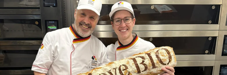 Ein Mann und eine Frau mit einem riesigen Ciabatta in der Hand. Auf diesem Steht "We love BIO". Beide lachen in die Kamera.